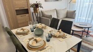 Spisebord dækket med tallerkener, skåle, glas og bestik, omgivet af stole og en sofa i et lysfyldt rum med en plante og et kunstværk på væggen.