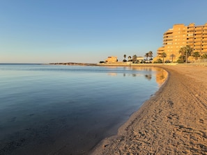 A beach with a calm sea, sandy shore, and a row of buildings in the background.