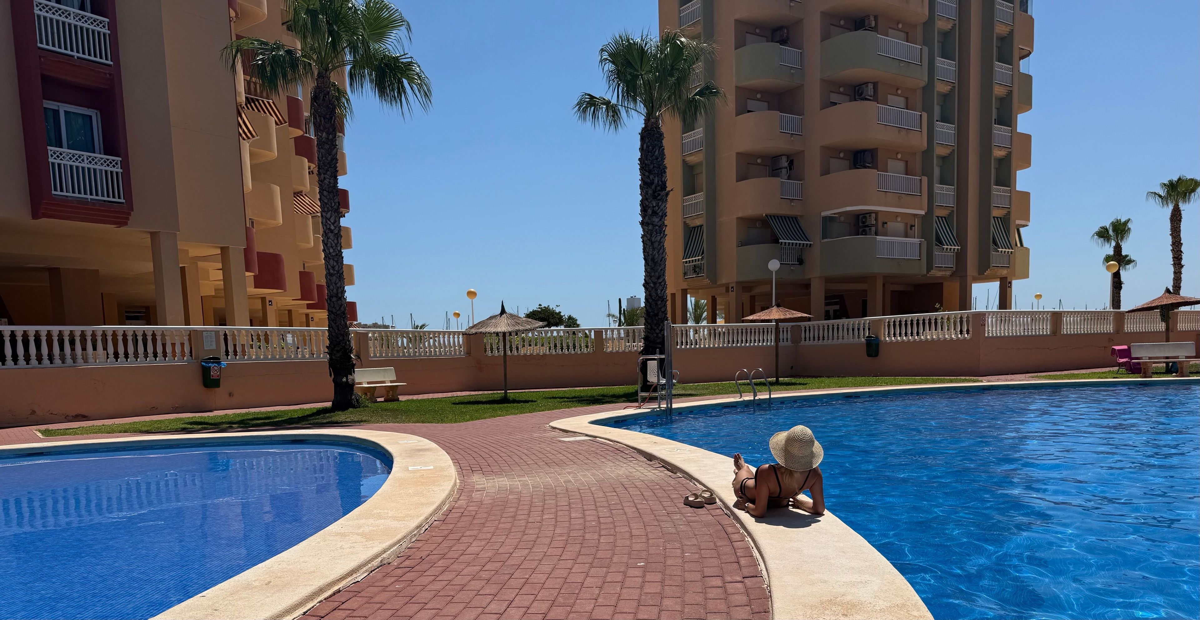 Una piscina con borde curvo, rodeada por un sendero de ladrillo, palmeras y un edificio de varios pisos con balcones.