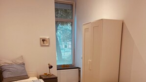 A bedroom with two beds, a wardrobe, a radiator, a window with blinds, a bedside table with a lamp, and a patterned rug. - Eli and Eli 1 Vacation Rental in the Heart of Berlin / Near Alexanderplatz (Berlin)