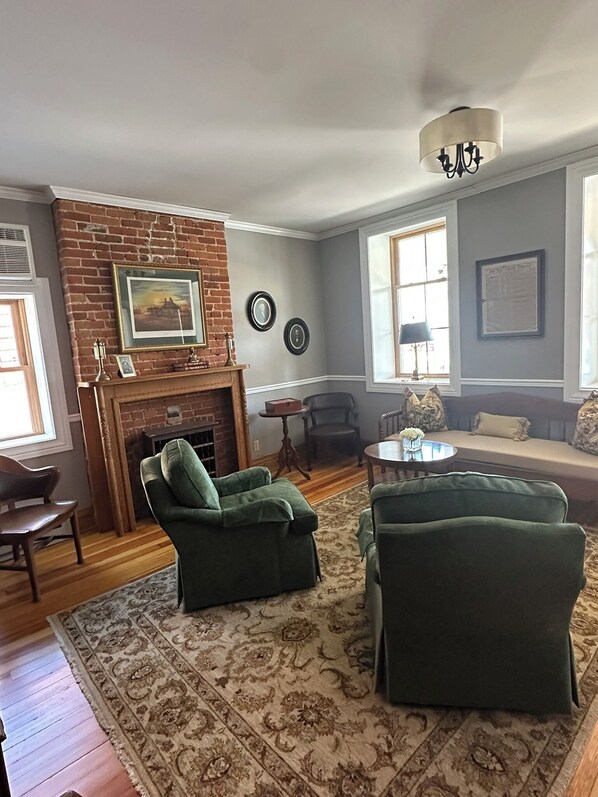A living room with a brick fireplace, green armchairs, a sofa, a round coffee table, and a chandelier.
