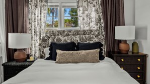 Ein Bett mit weißem Kopfteil, zwei Nachttische mit Lampen, ein brauner Teppich und ein Fenster mit Vorhängen.