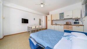 Quarto com cama, escrivaninha, cadeira, televisão e pequena cozinha.