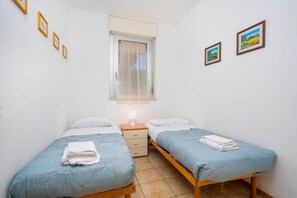 A bedroom with two beds, a window with a sheer curtain, a nightstand with a lamp, and framed pictures on the wall. - A comfortable and welcoming 61-square-meter apartment, perfect for six people, located on the ground floor (Scala A) of a building directly facing the seafront.The property is situated in Lignano Sabbiadoro in an excellent location, just a two-minute wal (Lignano Sabbiadoro)