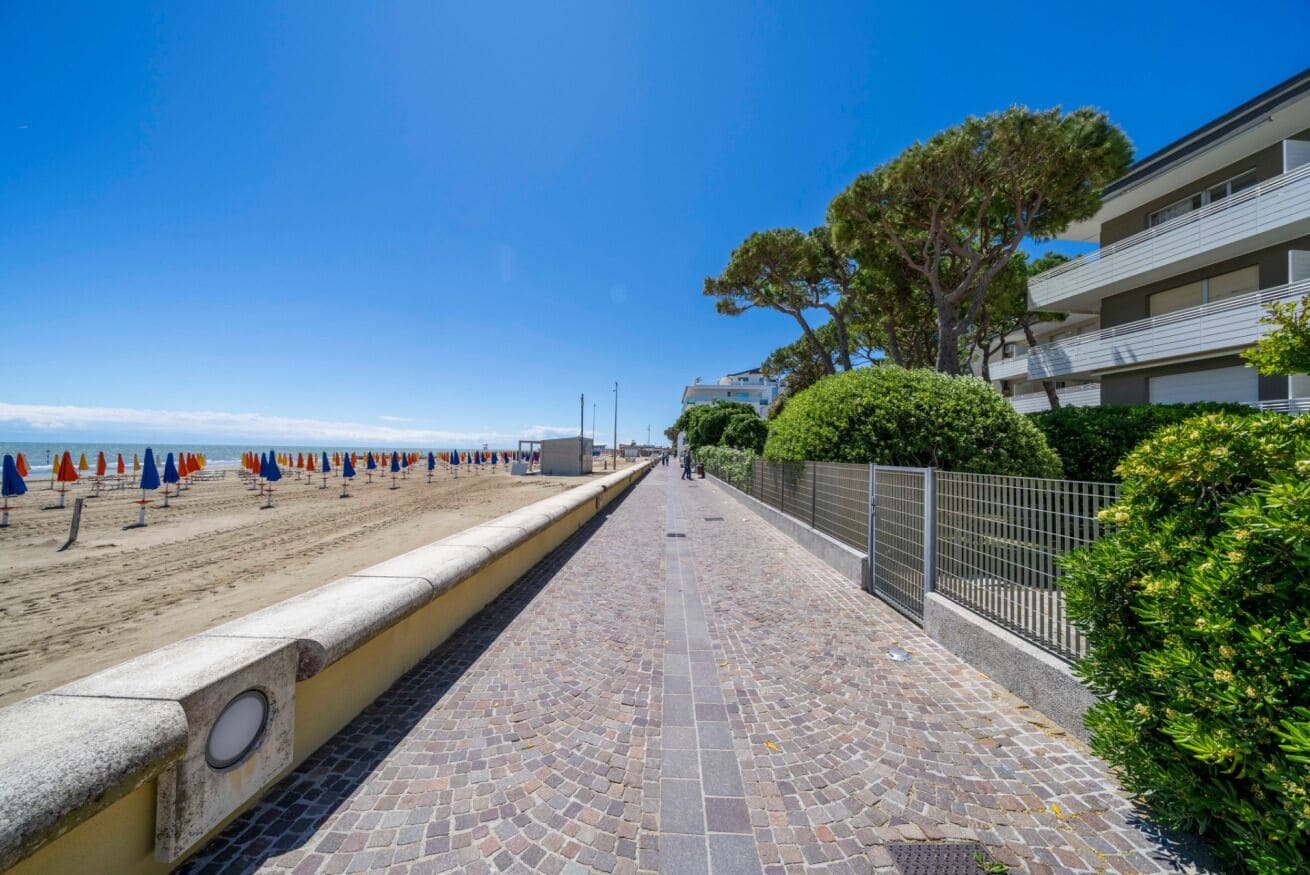 Ein gepflasterter Weg entlang eines Strandes mit Sonnenschirmen, einem Zaun und einem Gebäude mit Balkonen.