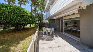 Um pátio com mesa e cadeiras, uma porta de vidro que dá para um espaço interior e um gramado bem cuidado com árvores.