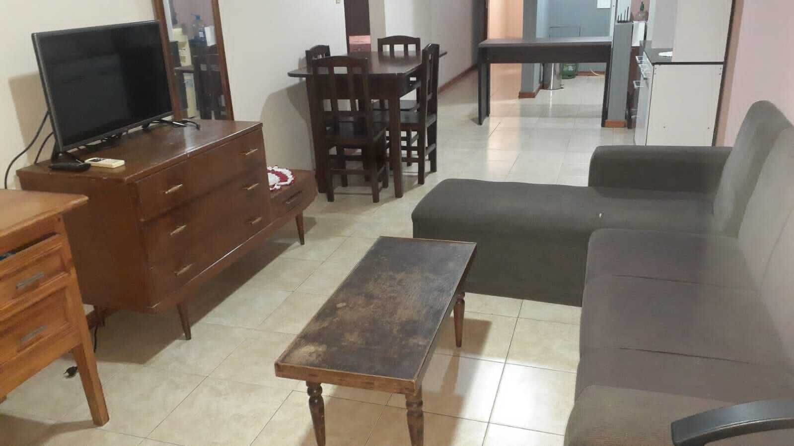 A living room with a sofa, a wooden coffee table, a TV on a wooden cabinet, a dining table with chairs, and a kitchen area in the background.