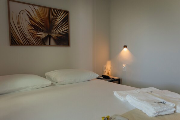 Habitación de hotel con una cama grande, almohadas blancas, una obra de arte colgada en la pared, una lámpara de noche y una toalla doblada con cuidado sobre la cama.