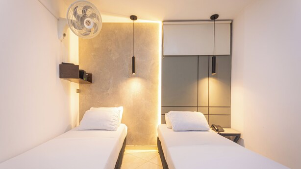 Une chambre d’hôtel avec deux lits, un ventilateur fixé au mur, une table de chevet avec un téléphone et une armoire avec un miroir.
