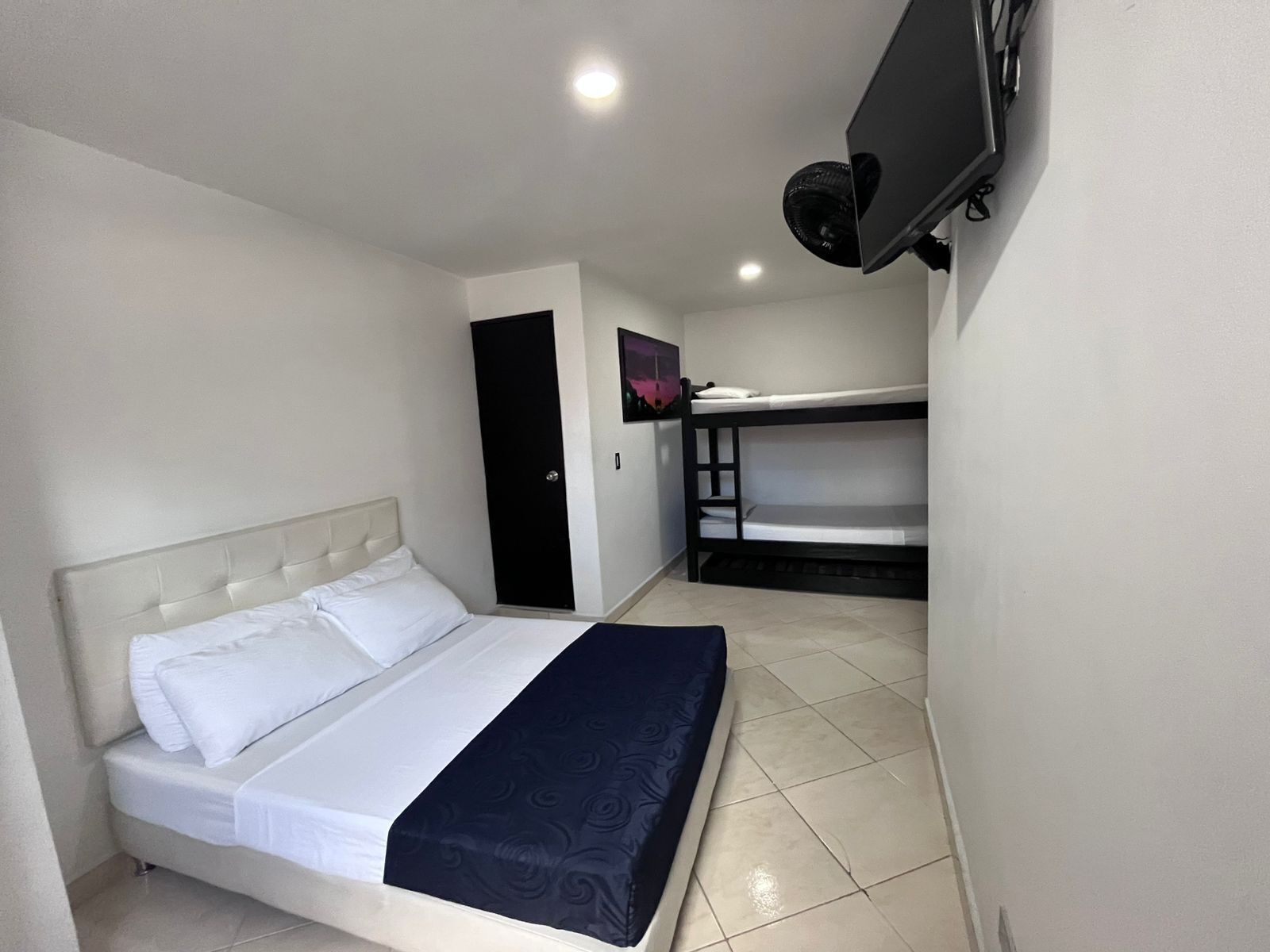 A room with a bed, a bunk bed, a door, a TV, and a wall-mounted fan.