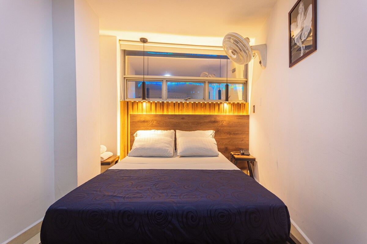 A hotel room with a bed, bedside table, lamp, and a window with a view of the cityscape.