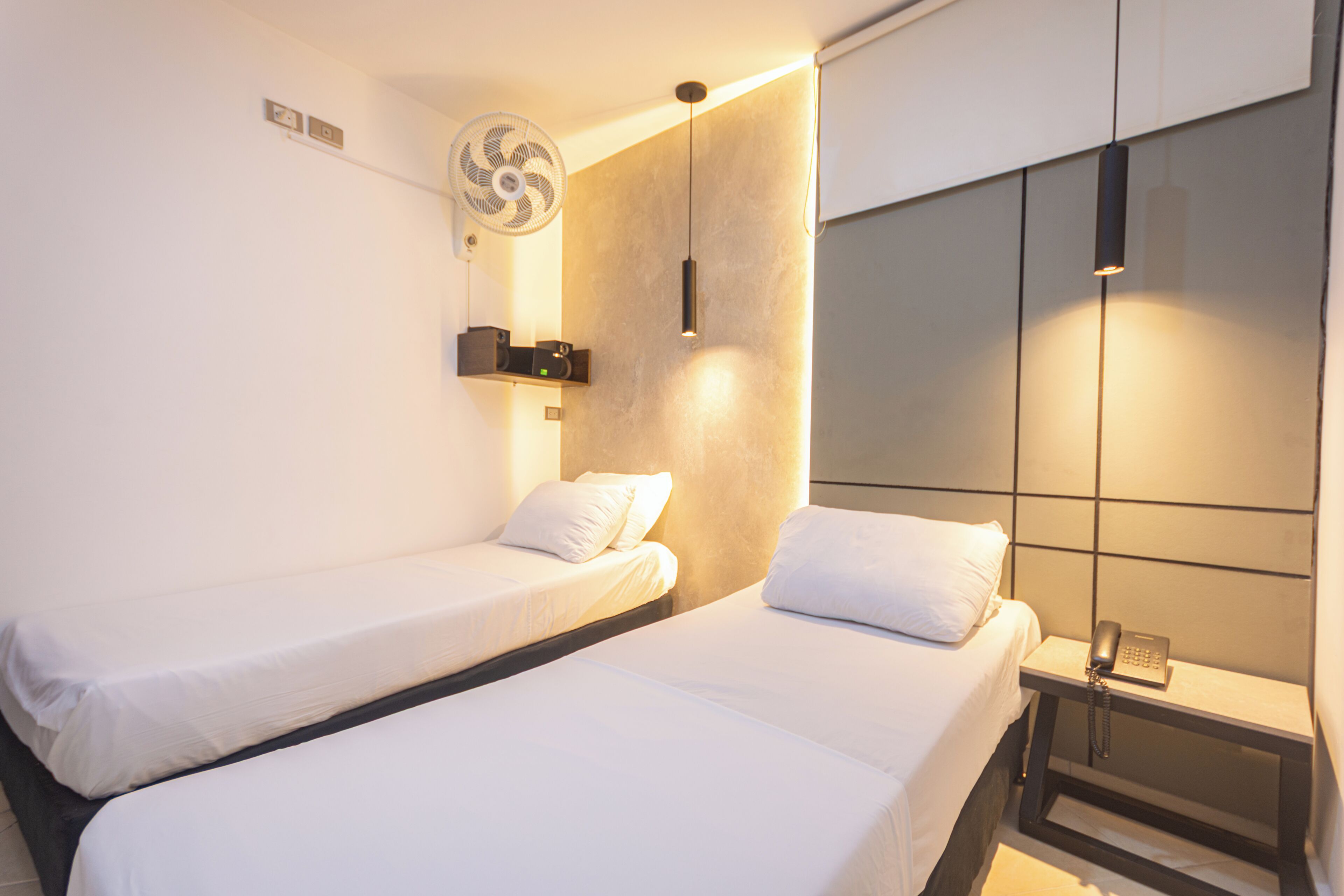 A hotel room with two beds, a bedside table with a phone, a wall-mounted fan, and a light fixture.