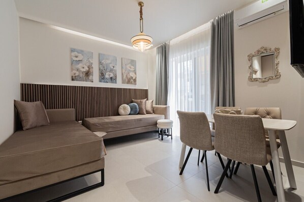 A modern living room with a sofa, a dining table with chairs, a small stool, and a television. - Marianthi Home by Rentals Pro - Nea Moudania Halkidiki (Nea Moudania)
