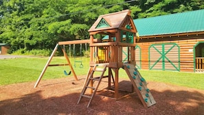 Une structure de jeux en bois, dotée d’un toboggan et de balançoires, qui se dresse sur fond de champ verdoyant et de cabane en rondins.