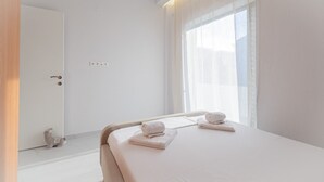A hotel room with a bed, two towels, a nightstand, a window with sheer curtains, and a door. - Dimitra Home by Rentals Pro - Polychrono Halkidiki (Polychrono)