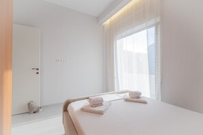 Ein Hotelzimmer mit einem Bett, zwei Handtüchern, einem Nachttisch, einem Fenster mit transparenten Vorhängen und einer Tür.