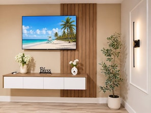 Una sala de estar con un televisor instalado en la pared, una consola de madera con un florero y una planta en maceta, además de una lámpara fijada a la pared.