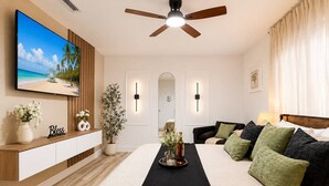 Un dormitorio con cama, televisor, ventilador de techo, mesita de noche, planta y televisor de pared mostrando una escena de playa.