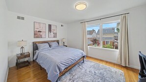 Une chambre à coucher avec un lit, des tables de chevet, une fenêtre avec des rideaux et des œuvres d’art accrochées au mur.