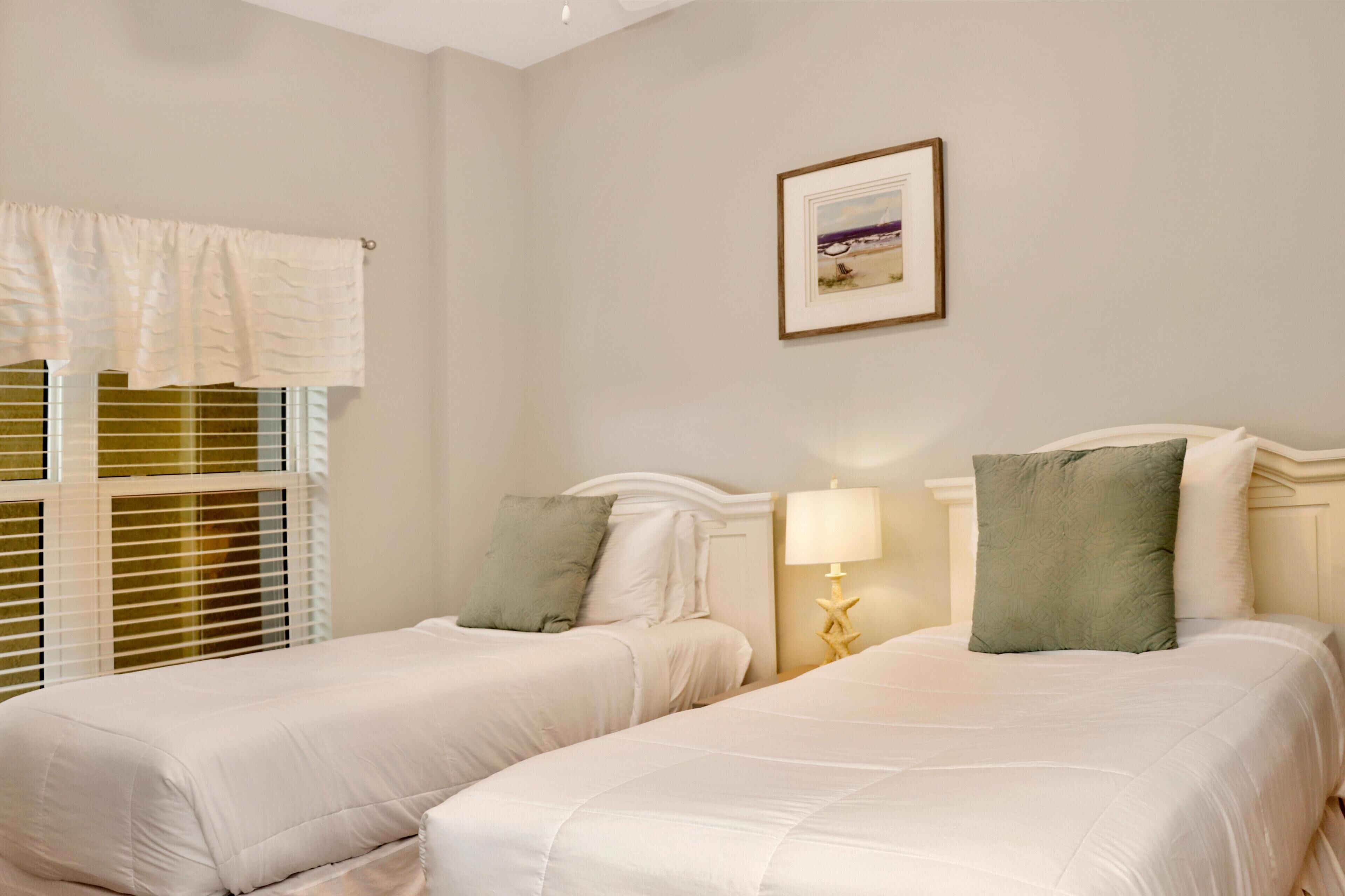 A hotel room with two beds, a window with blinds, a framed picture on the wall, and a nightstand with a lamp.
