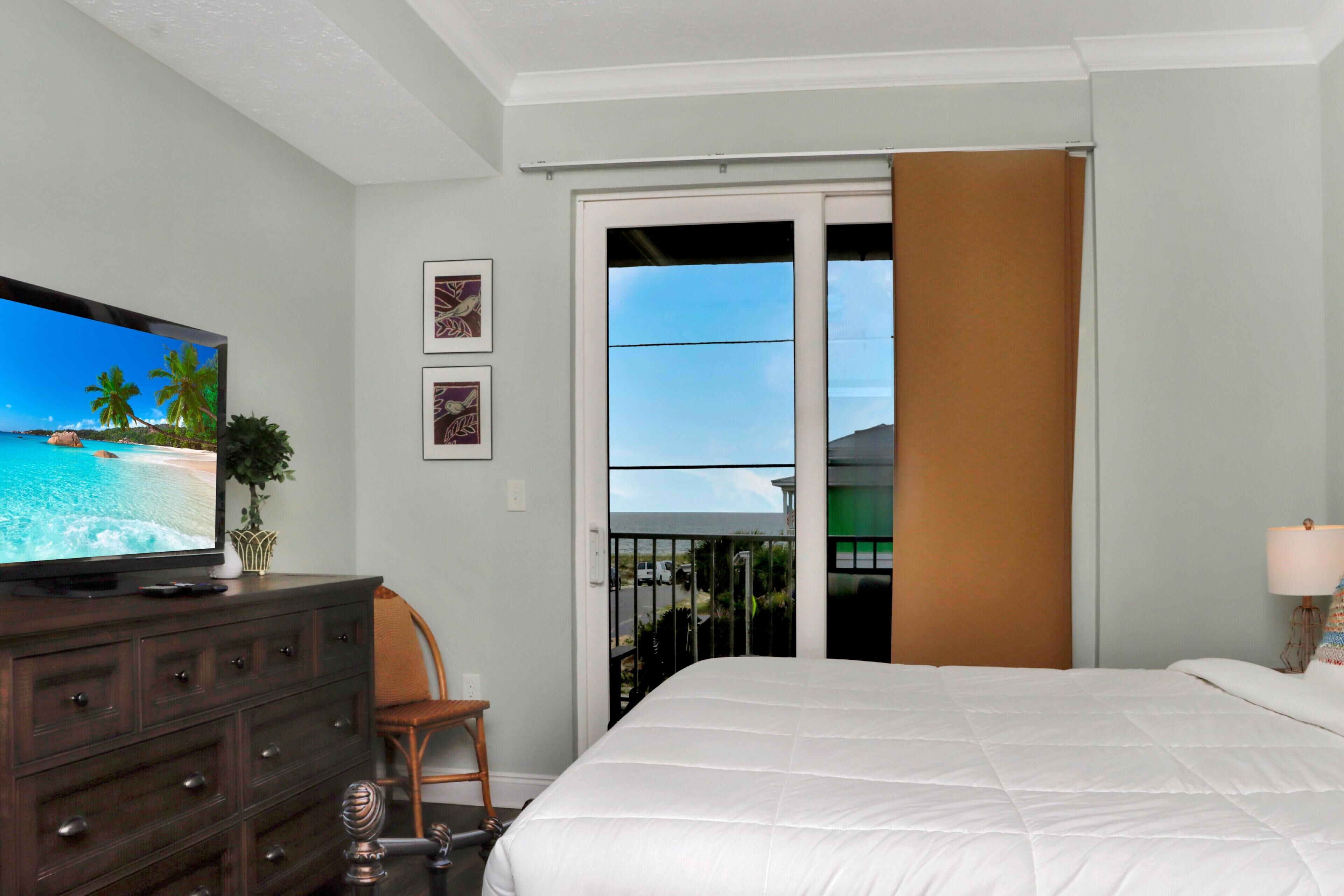 Ein Hotelzimmer mit einem Bett, einem Fernseher, der eine Strandansicht zeigt, einem Schminktisch, einem Sessel und einer Schiebetür zu einem Außenbereich.