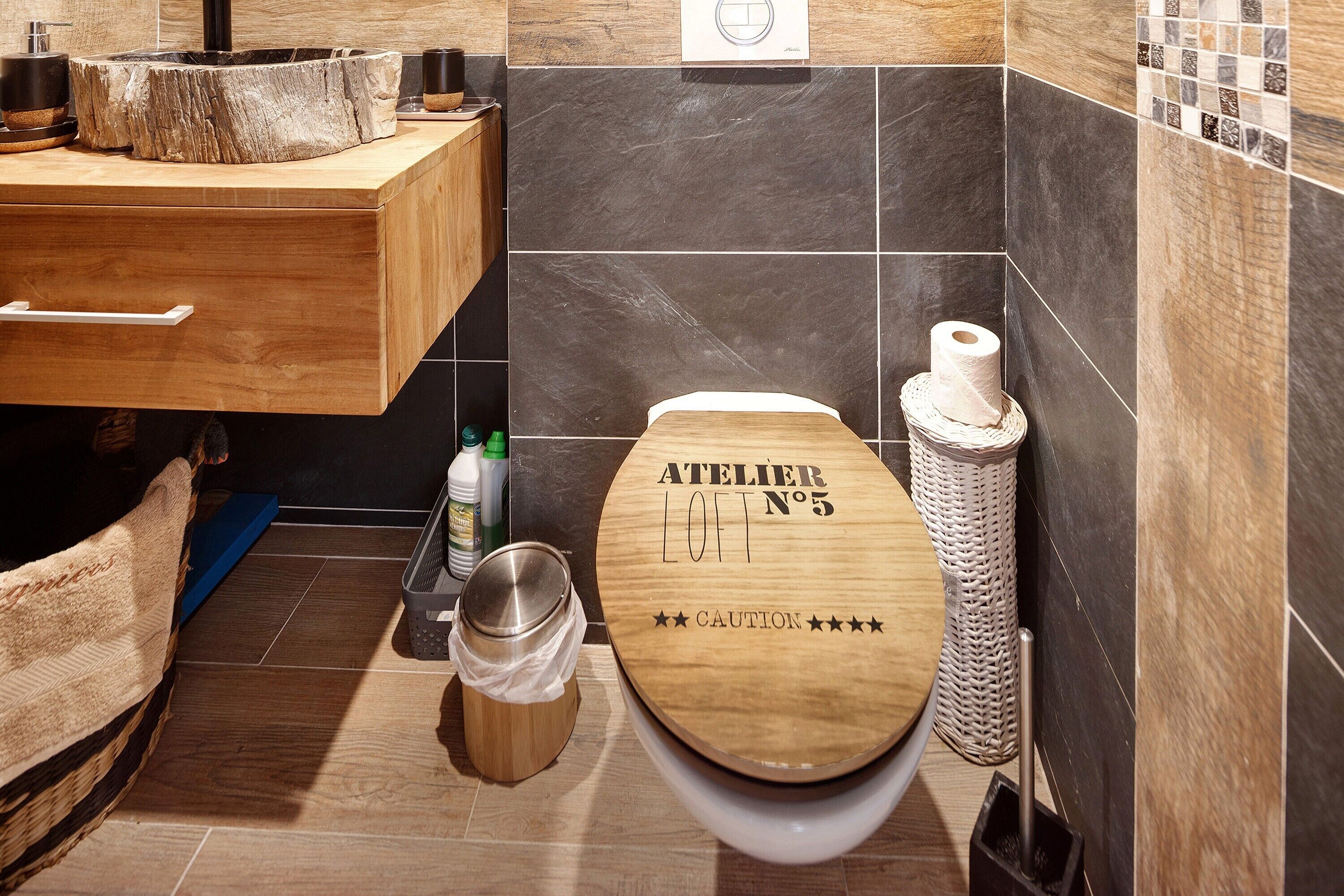 A bathroom with a wooden toilet, a sink with a wooden countertop, a trash bin, and a toilet brush.