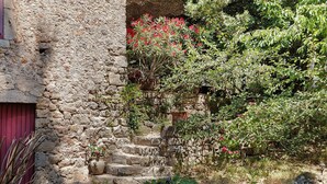 Ein Steinhaus mit roter Tür, einer Treppe, die zu einer Holztür führt, und einem Garten mit verschiedenen Pflanzen und Blumen.