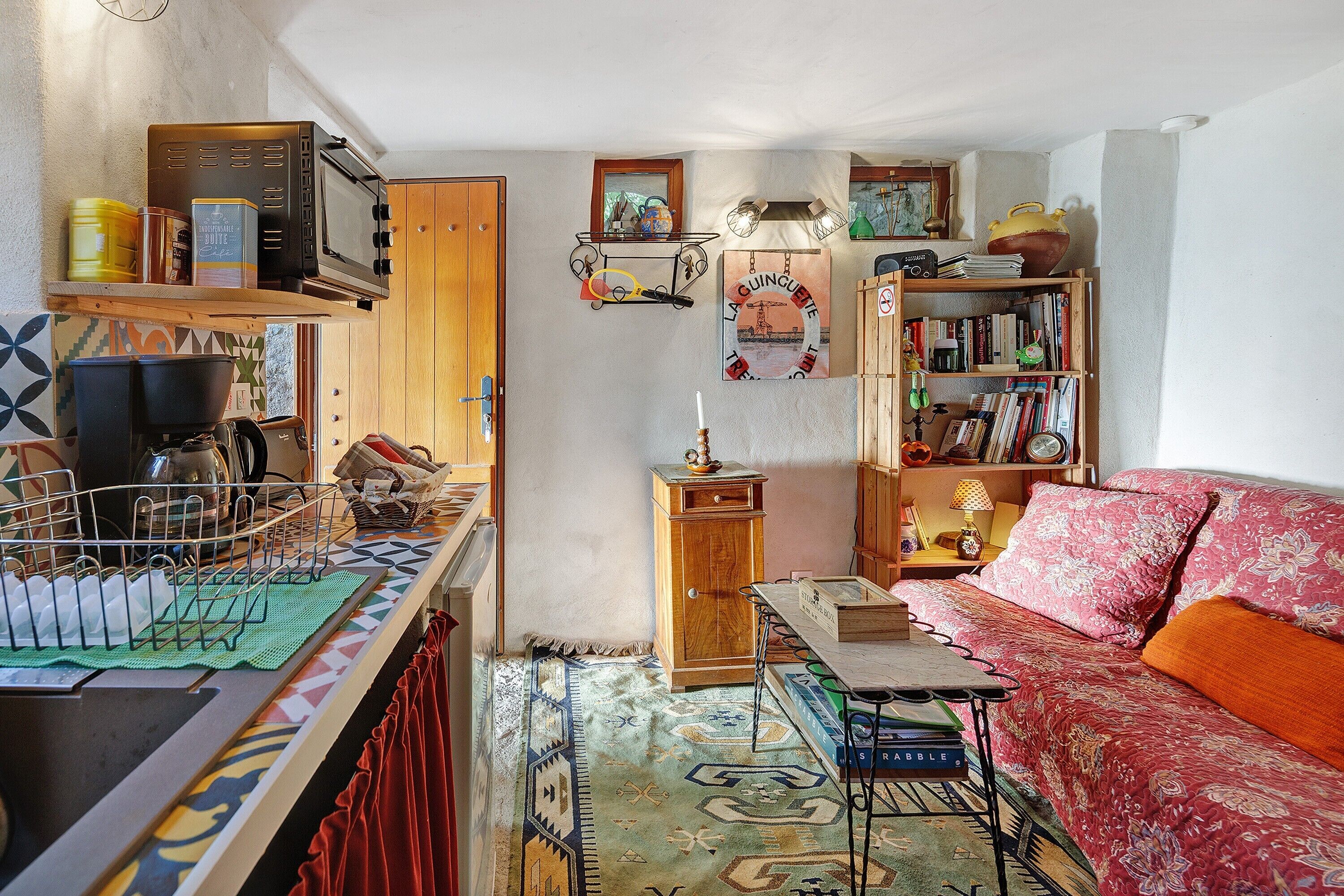 Un espace de vie compact comprenant un coin cuisine, une bibliothèque, un tapis à motifs et un canapé recouvert d’un tissu à fleurs.