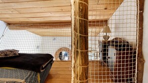 Eine Holzbalkendecke, ein Bett mit einem gemusterten Überwurf, ein Moskitonetz und ein kleines, rundes Fenster.