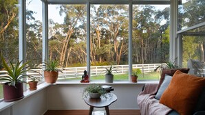 A room with a large window, a sofa, a small table, and several potted plants. - Hepburn Woodland Retreat (Dry Diggings)