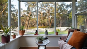 A room with a large window, a sofa, a small table, and several potted plants. - Hepburn Woodland Retreat (Dry Diggings)