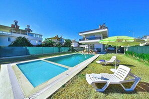 A swimming pool with lounge chairs and an umbrella, surrounded by a fence and buildings. - Villa Esma - 3 Bed with private garden (Antalya)