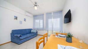 Ein Wohnzimmer mit einem blauen Sofa, einem Holzespeisetisch, Stühlen, einem Deckenventilator und einem an der Wand befestigten Fernseher.