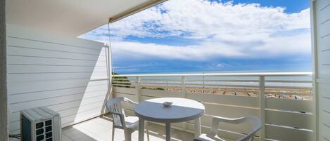 A balcony with a view of the beach, a table, two chairs, and an air conditioning unit.