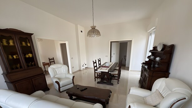A spacious living room with a dining area, white leather sofas, a wooden dining table, and a chandelier. - ‘Casa Vacanze Rita La California’ Apartment with Private Garden and Balcony (La California)