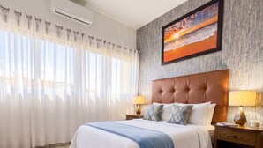 Ein Hotelzimmer mit einem großen Bett, einem Deckenventilator, einer Klimaanlage, einem Nachttisch mit Lampe und einem Gemälde an der Wand.