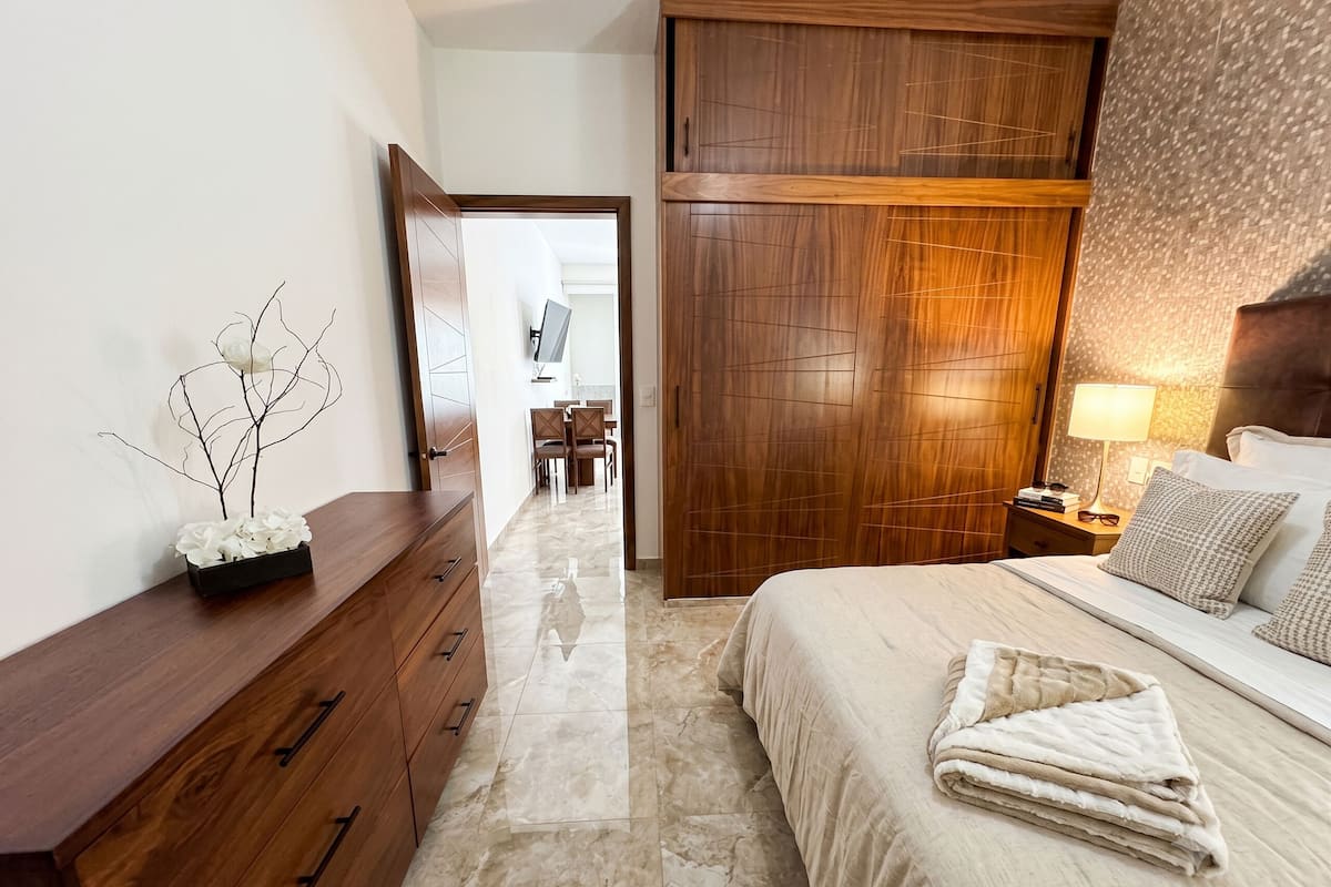 Quarto de hotel com uma cama, cômoda de madeira, mesa de cabeceira com abajur e vista para uma sala de jantar através de uma porta aberta.