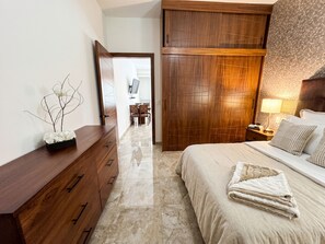 Habitación de hotel con cama, cómoda de madera, mesita de noche con lámpara y vista a un comedor por una puerta abierta.