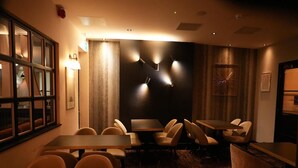 A dimly lit room with tables and chairs, a patterned carpet, and a large window with blinds. - Deluxe Double Room (Derby)