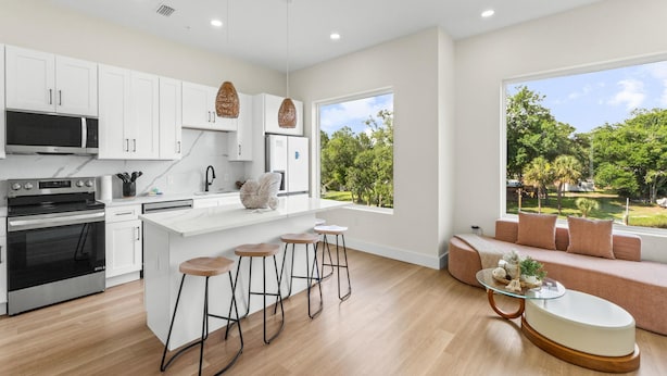 A modern kitchen with white cabinets, a central island with stools, a dining area with a glass table, and a large window offering a view of a green landscape. - 3008 Whispering Sand C (Panama City Beach)