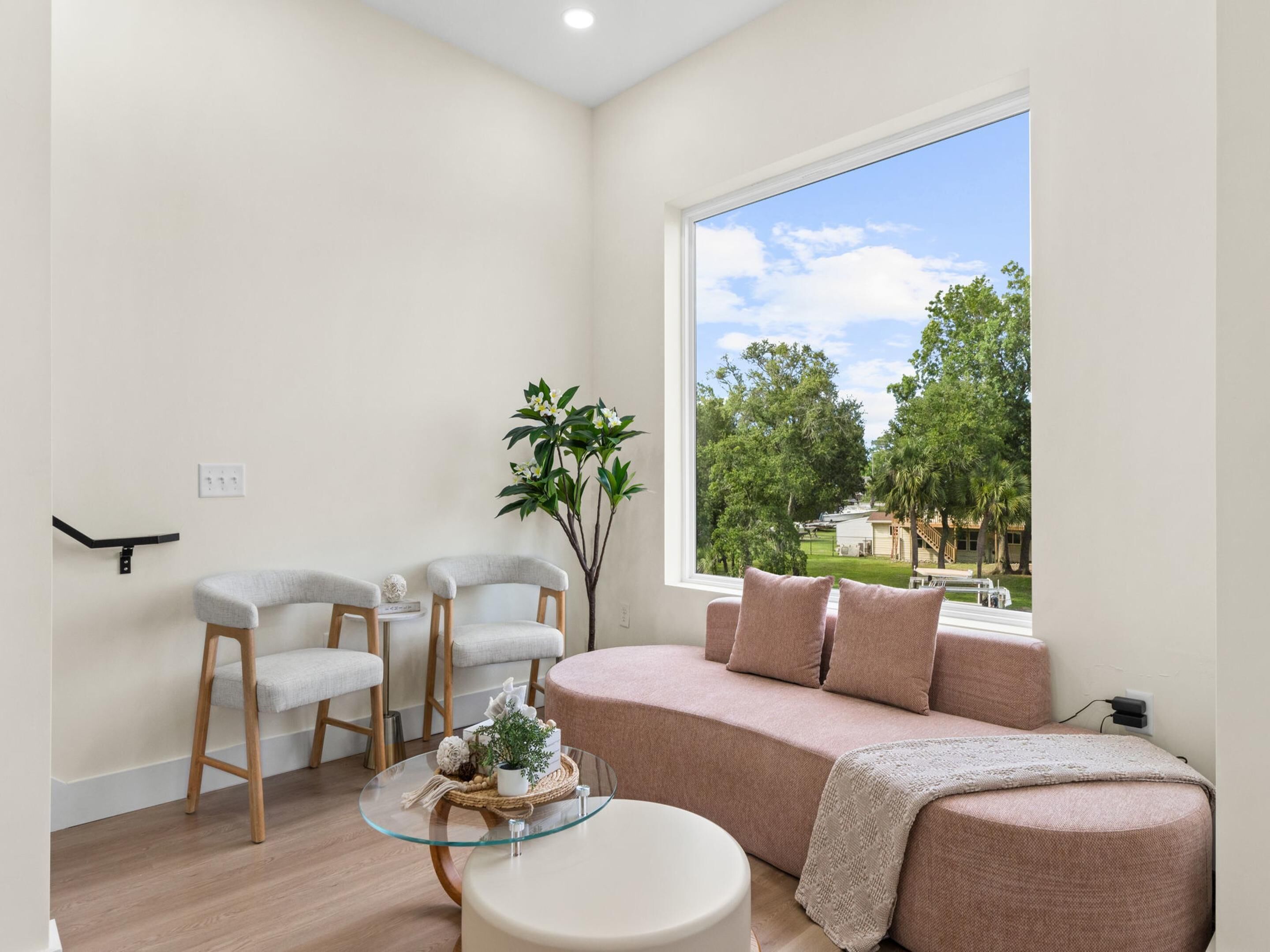 Una sala de estar moderna con un sofá, dos sillones, una mesa de centro de vidrio y un ventanal con vistas a árboles y una casa.