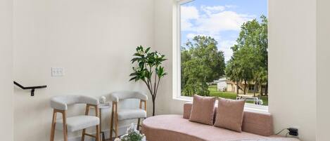 Una sala de estar moderna con un sofá, dos sillones, una mesa de centro de vidrio y un ventanal con vistas a árboles y una casa.