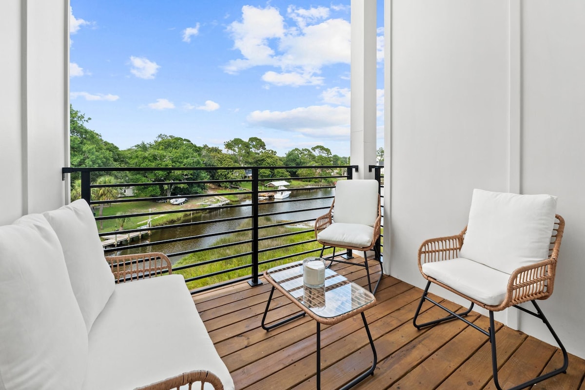 A balcony with wicker chairs, a glass table, and a view of a river and greenery.