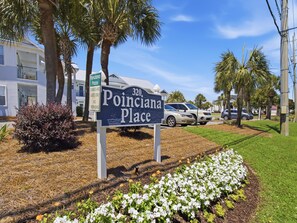 A sign for 'Poinciana Place' with a clear view of the surrounding area including a building, parked cars, and a landscaped garden. - Poinciana Place #222 - Mystic Miramar (Miramar Beach)