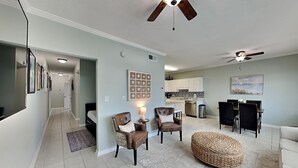 A spacious living area with a dining table, two armchairs, a woven ottoman, ceiling fans, and a kitchen in the background. - Poinciana Place #222 - Mystic Miramar (Miramar Beach)