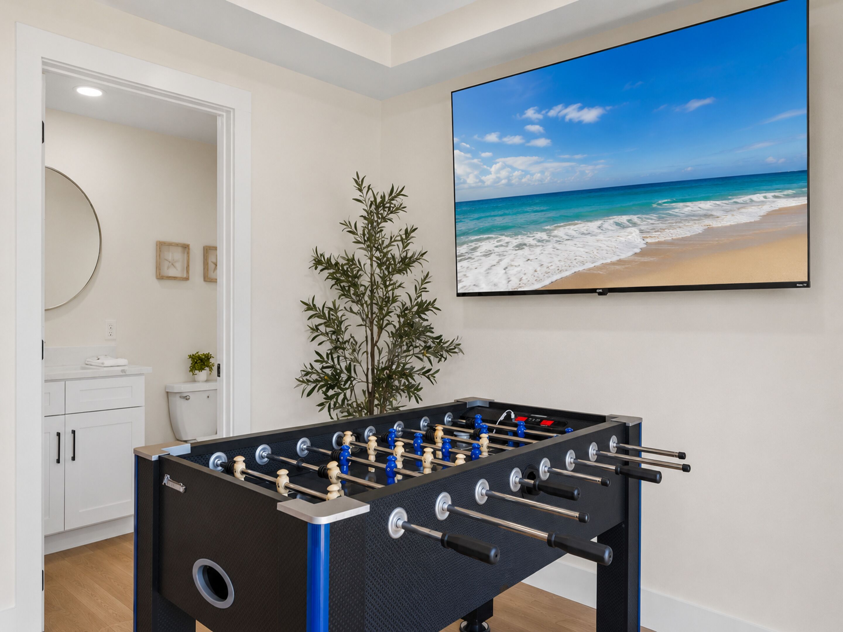 A room with a foosball table, a television displaying a beach scene, a plant, and a bathroom with a toilet and sink.