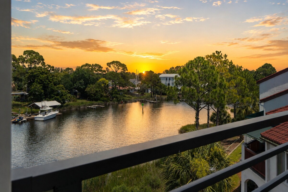 Vista del atardecer desde un balcón con vistas a un lago, con barcos, árboles y casas.