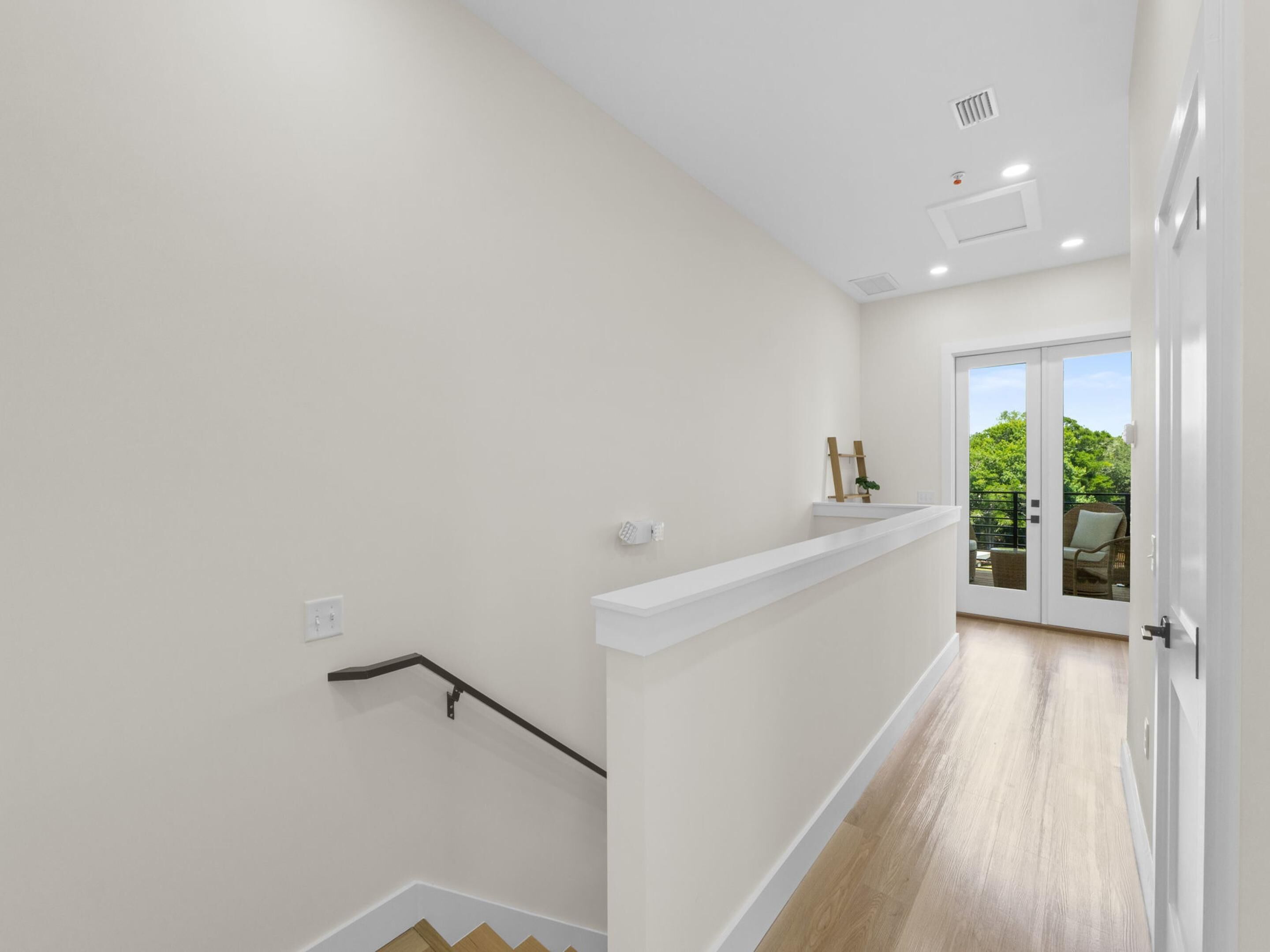 A staircase with a handrail, a white wall, and a door leading to a balcony with a view of greenery.