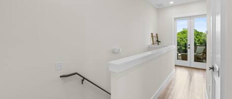 A staircase with a handrail, a white wall, and a door leading to a balcony with a view of greenery.