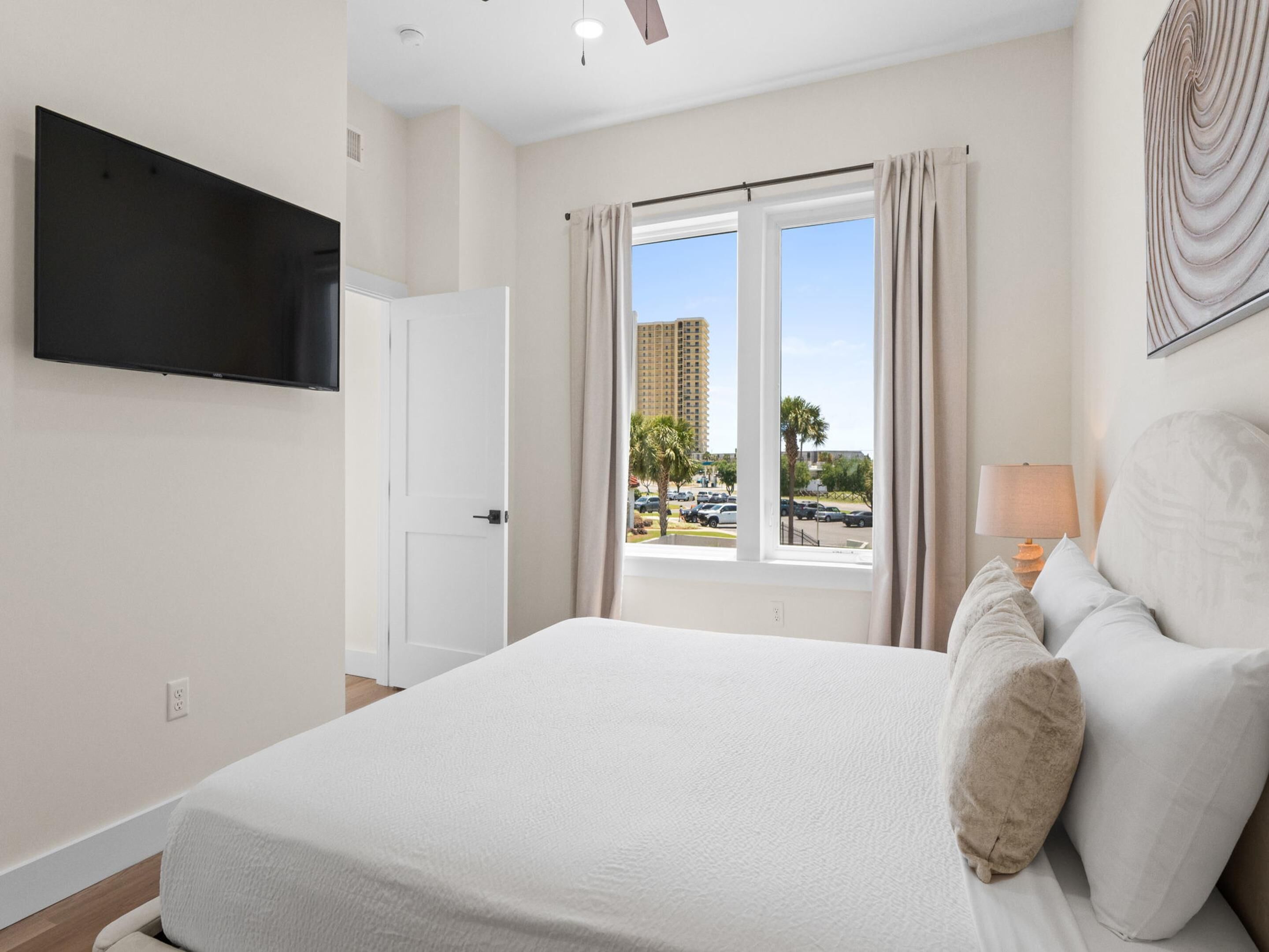 A bedroom with a bed, a TV mounted on the wall, a window with curtains, and a view of buildings and cars outside.